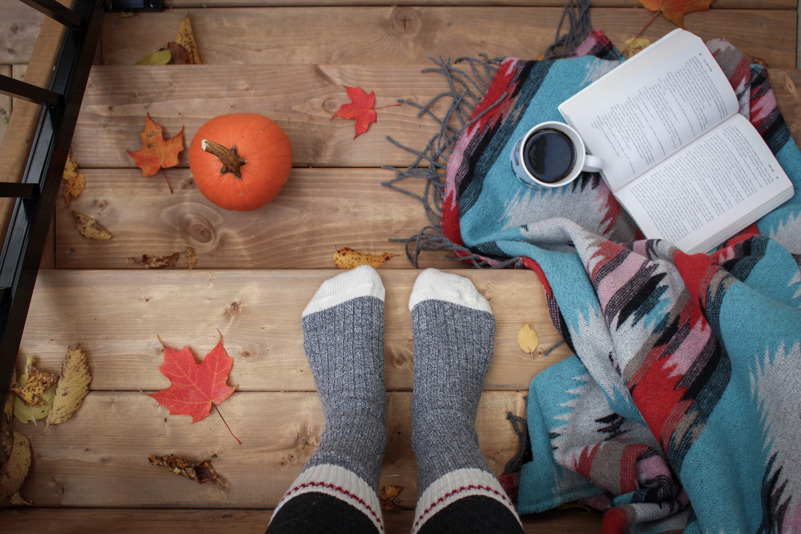 Picture of feet with socks with leaves, a pumpkin, coffee, soutwest blanket and a book.